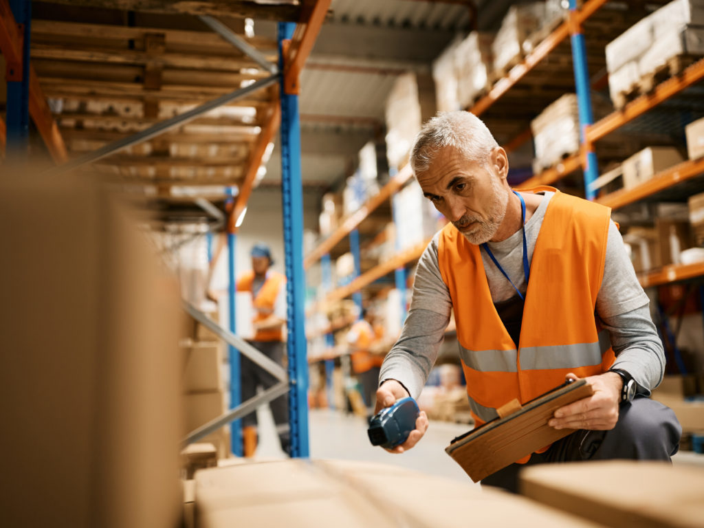 Mature warehouse worker using touchpad and scanning cardboard boxes at storage compartment.